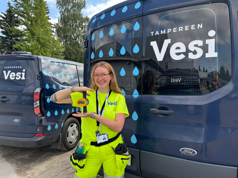 Arnika Tikka pitämässä etäluettavaa vesimittaria kädessä, Tampereen Veden autojen edessä.